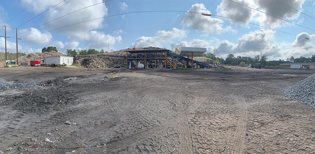 Daytime image wide filed view of debris pile, behind dirt area cris-crossed with large tire tracks, a conveyor belt covered by sturdy aluminum cover with flourescent lighting
