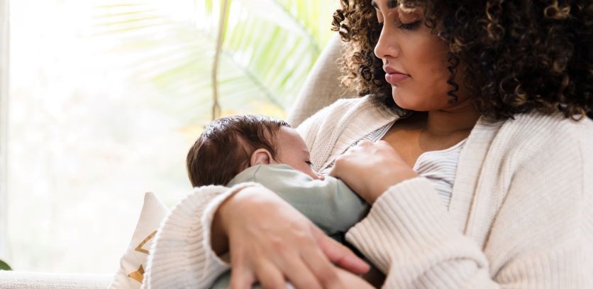 Baby boy in sage green onesie breasfeeding with his mom, an African American woman with long, dark curly hair who is wearing a beige cardigan