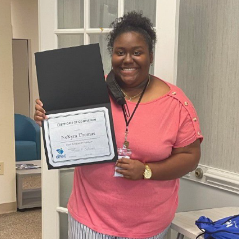 An African American young lady wearing a pink tee shirt and blue striped pants holds up her certificate of internship completion.