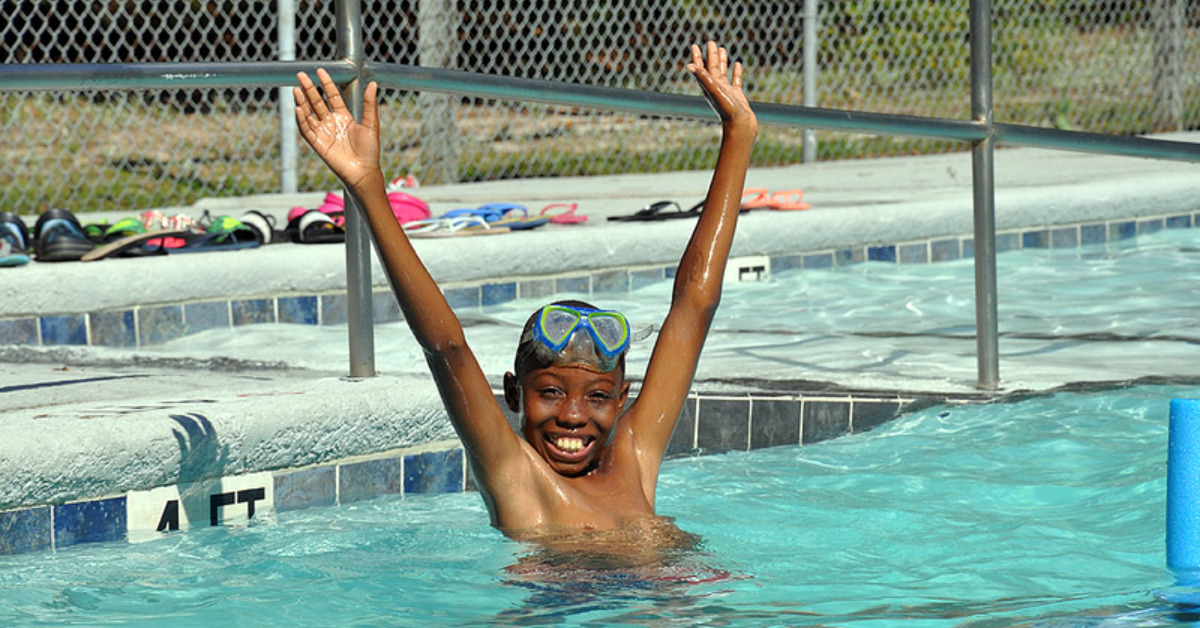 African American boy in pool with arms raised up, cheering, while smiling at the camera. He has blue swimming goggles on his forehead.
