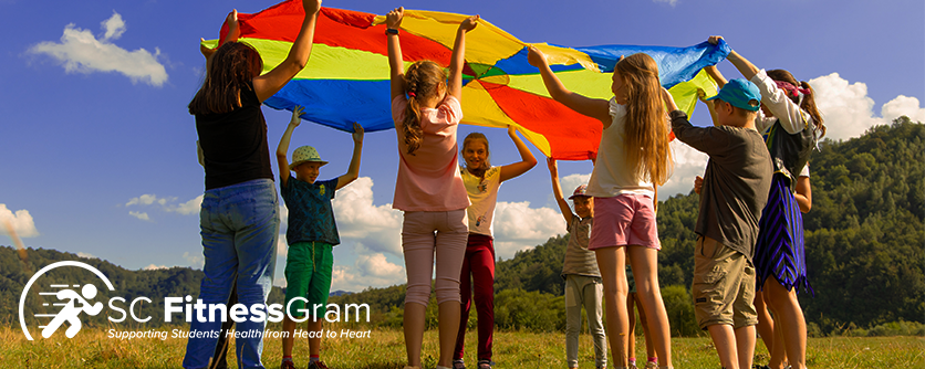 Group of children in a circle raising a rainbow sheet over their heads. The sky is blue with a few clouds. The SC Fitnessgram logo is in all white in the bottom left corner.