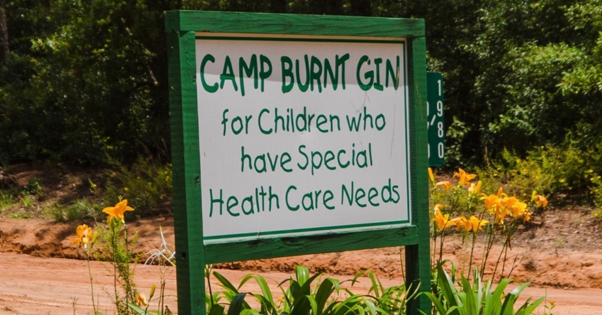 Wooden sign with green border and white paint is near the edge of a dirt road. In green paint, the sign reads "Camp Burnt Gin for Children who have Special Health Care Needs".
