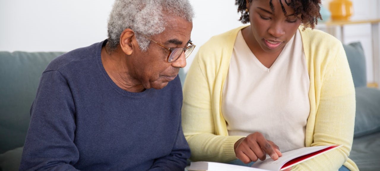 African American granddaughter wearing a white tee shirt and light yellow cardigan is reading to her grandfather, who has gray hair, a blue sweater, and glasses.