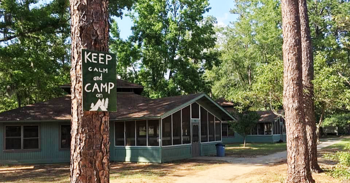 Looking down a dirt path with trees and blue cabins. The closest tree has a green sign handpainted with white letters saying "Keep Calm and Camp On".