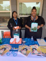 Two African American ladies wearing black tops with blue DHEC lanyards hold up displays from their COVID DHEC table 